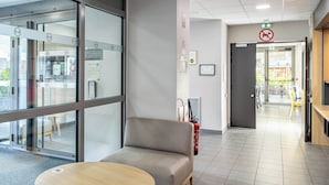 Un área de espera moderna con un sofá y una mesa redonda, una puerta de cristal de entrada y un extintor en la pared.