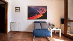 Ett rum med en blå soffa, en inramad bild av ett vulkanutbrott, ett element och en tv som står på ett skåp.