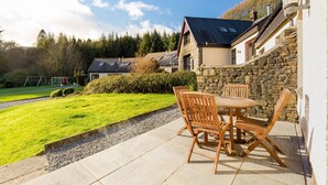 Un patio con una mesa y sillas, un muro de piedra y una casa con techo inclinado al fondo.