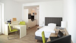 A modern hotel room with a bed, desk, and chair, a kitchenette, and a bathroom visible through an open door. - ADAPT APARTMENT HOTEL Giessen (Giessen)