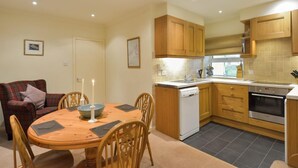 A compact kitchen with wooden cabinets, a dishwasher, and an oven. There is a dining table with chairs and a candle on it. - Weighbridge Cottage - a detached cottage with open fire and private garden (OBAN)