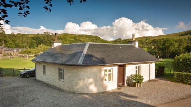 A small, single-story house with a sloped roof, a chimney, and a gravel driveway. - Weighbridge Cottage - a detached cottage with open fire and private garden (OBAN)