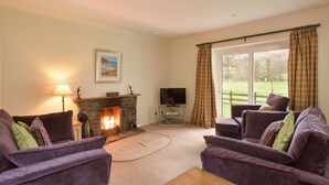 A living room with a stone fireplace, two purple sofas, a television, and a large window with curtains. - Weighbridge Cottage - a detached cottage with open fire and private garden (OBAN)