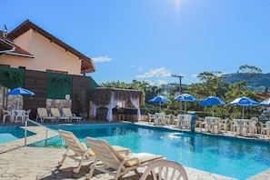 A swimming pool with lounge chairs and umbrellas, a building with a stone facade, and a view of hills in the background. - Termas Park Hotel (Gravatal)