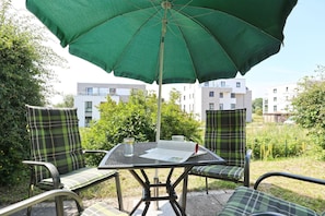 Ulkoterassi, jossa on pöytä ja tuoleja vihreän aurinkovarjon alla. Alue on vehreän kasvillisuuden ympäröimä, ja taustalla näkyy moderneja rakennuksia.