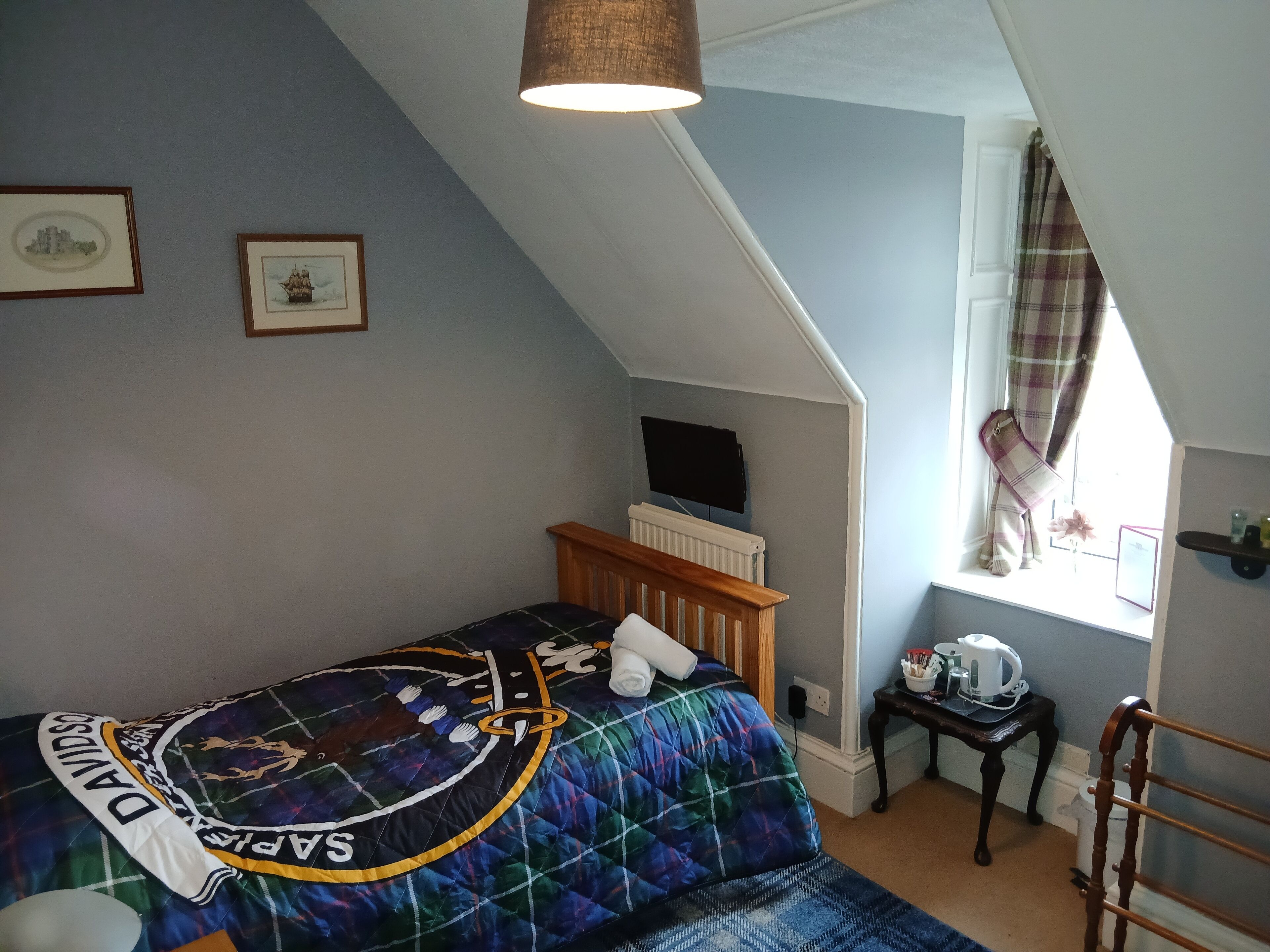 A bedroom with a bed, a window with curtains, a radiator, and a small table with a kettle.