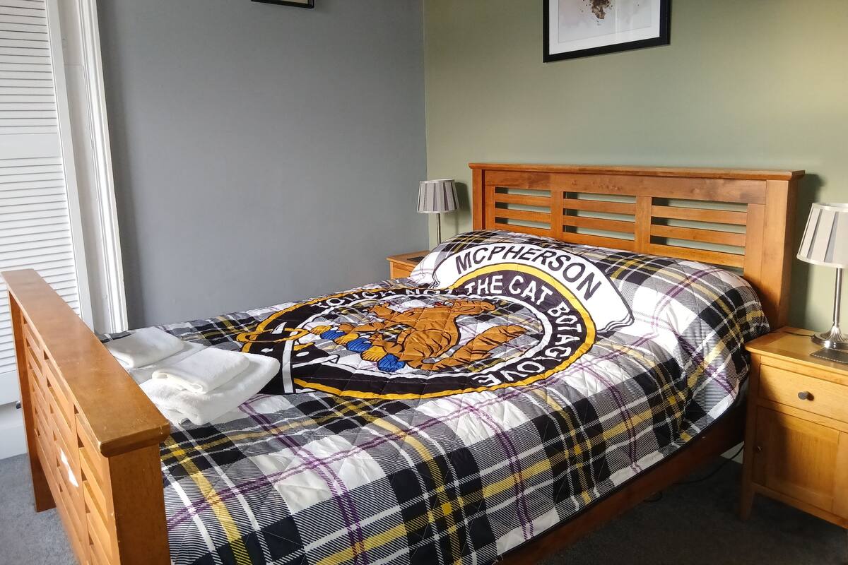 A bedroom with a plaid bedspread, a wooden headboard, a nightstand with a lamp, and a framed deer picture on the wall.