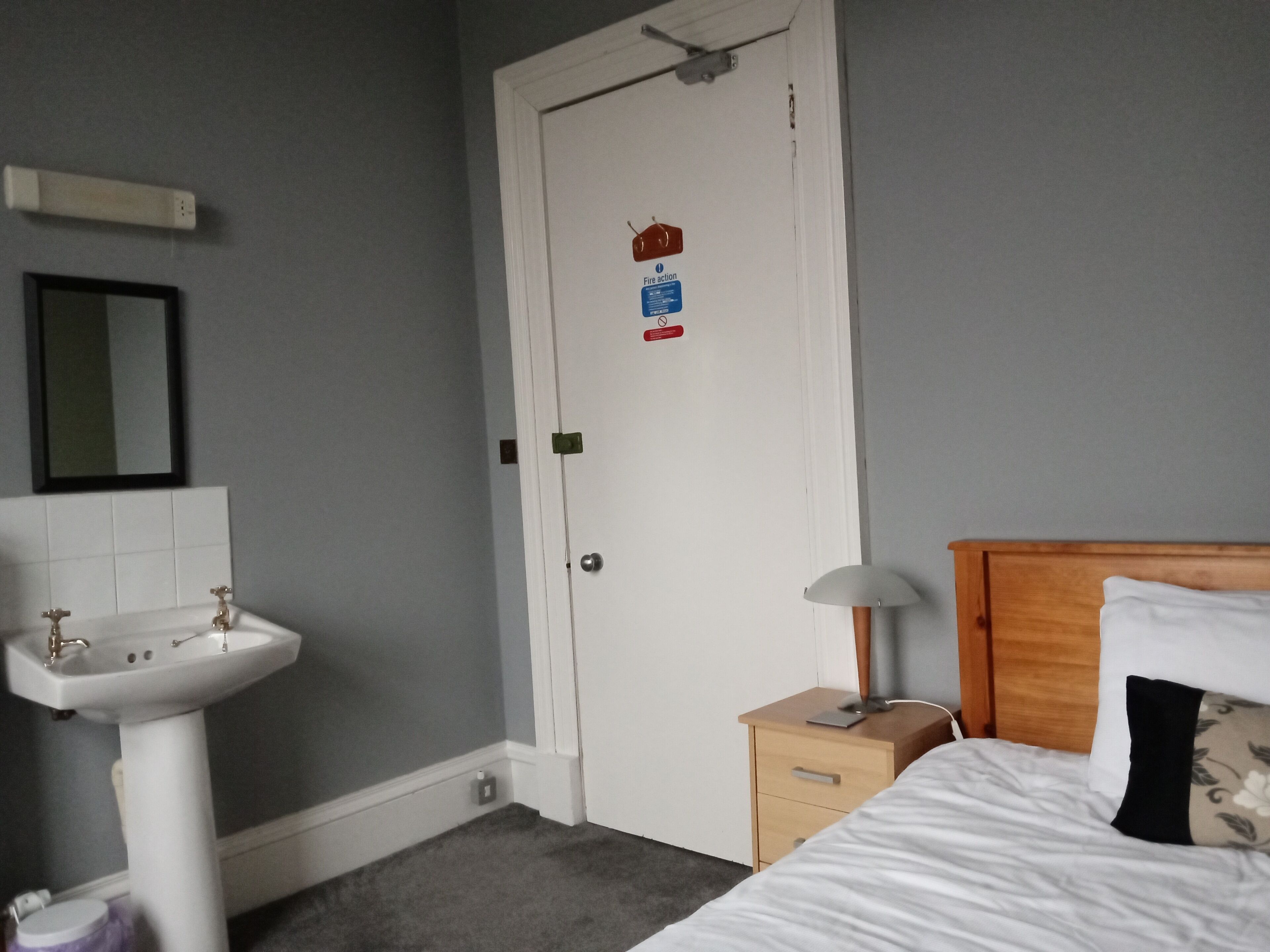 Un dormitorio con cama, mesita de noche con lámpara, lavamanos blanco y una puerta con señal de evacuación contra incendios.