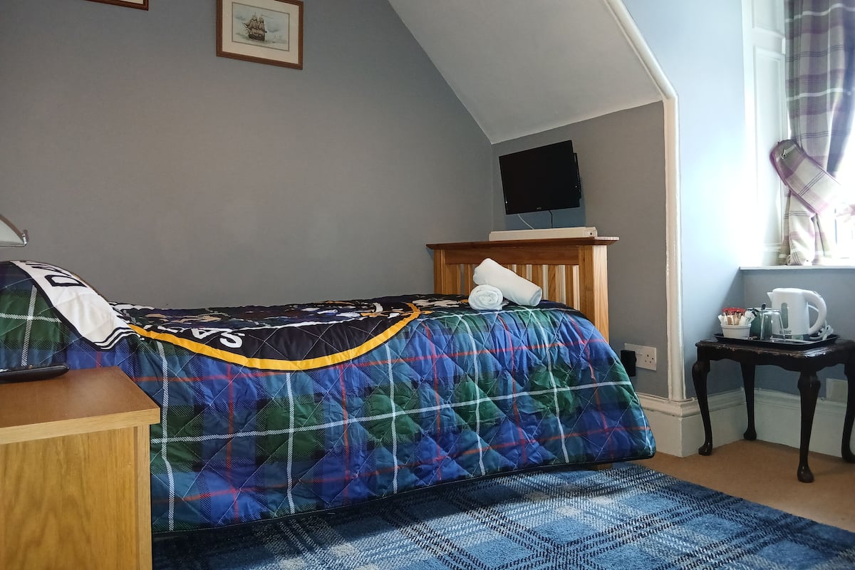 A bedroom with a bed, a wooden nightstand, a small table with a kettle, and a television on the wall.