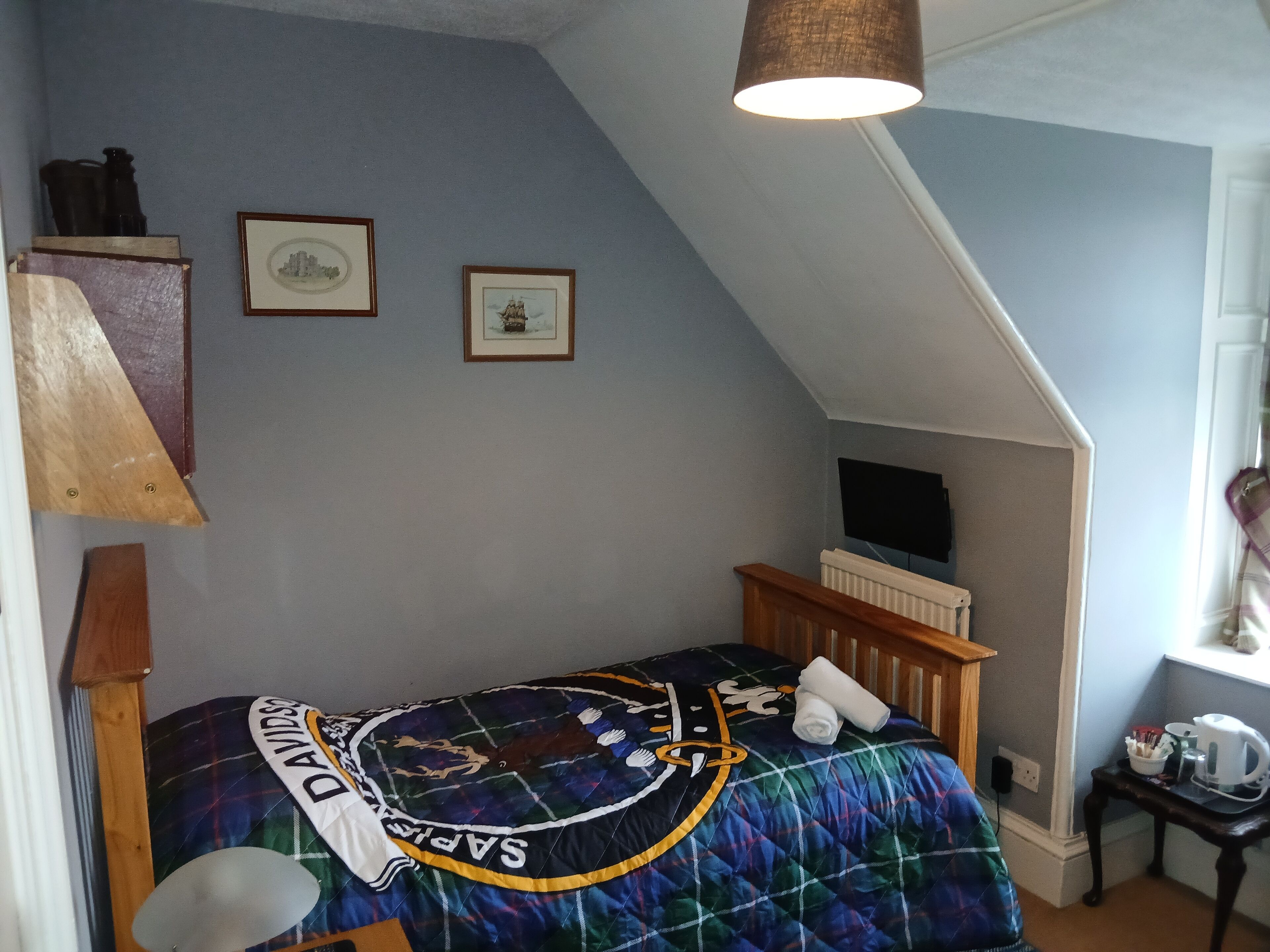 A bedroom with a sloped ceiling, a bed with a quilted blanket, two framed pictures on the wall, a wooden nightstand, and a lamp.