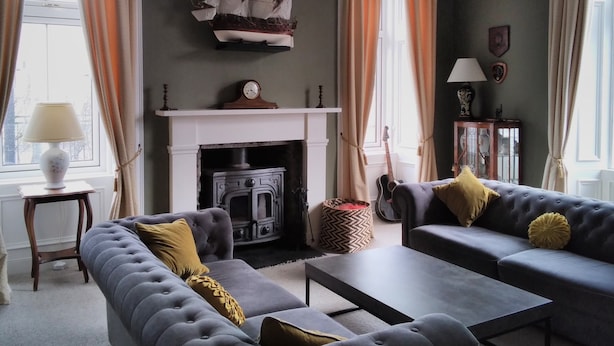 A living room with a fireplace, two sofas, a table, a guitar, and a ship decoration on the wall. - Greenland House B&B (Castletown)