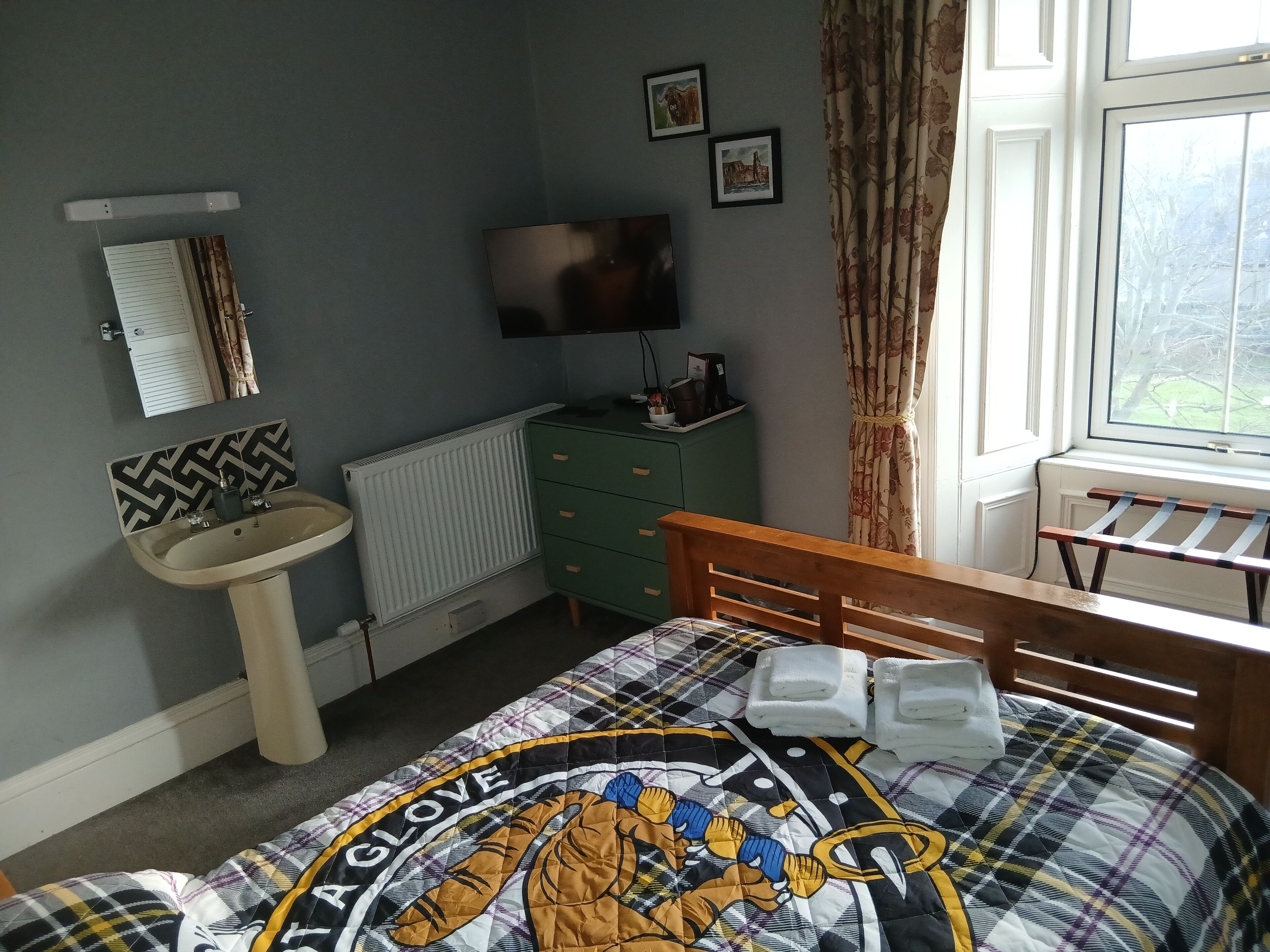 A bedroom with a bed, a bedside table, a dresser, a radiator, a window with curtains, and a wall-mounted TV.