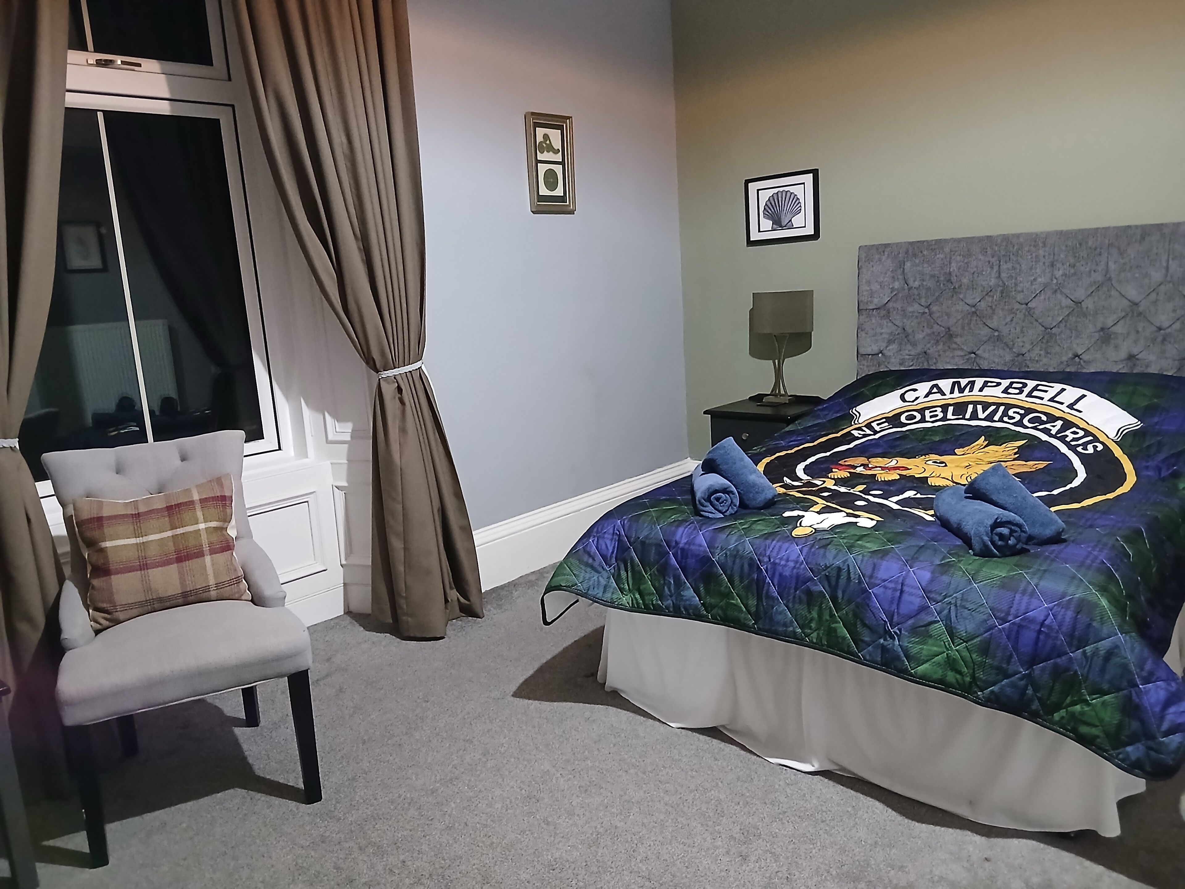 Un dormitorio con una cama con un edredón colorido, una silla con un cojín escocés, una ventana con cortinas y una mesita de noche con una lámpara.