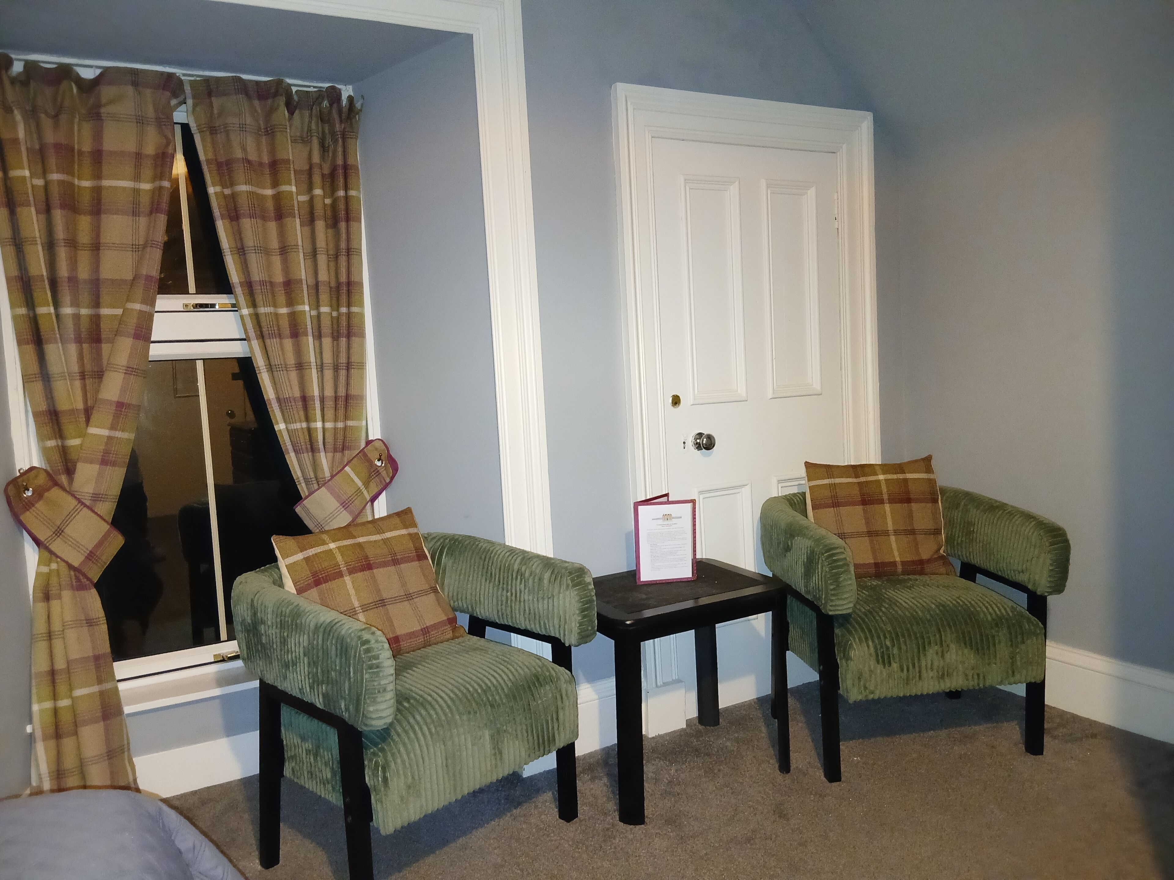 A room with two green armchairs, a small table, a white door, and plaid curtains.