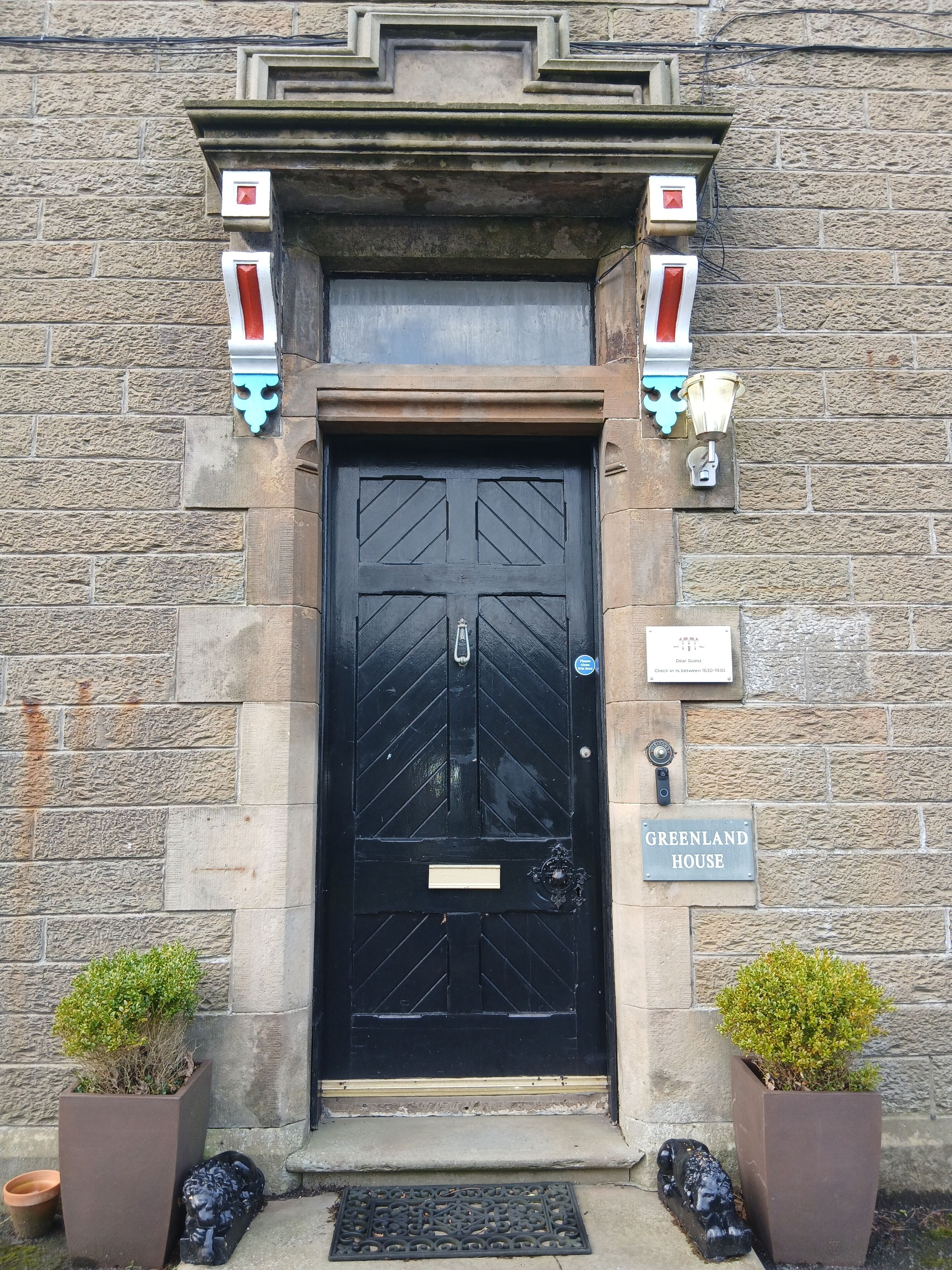 A black door with a diamond pattern, flanked by stone walls and two potted plants. A sign above the door reads "GREENLAND HOUSE".
