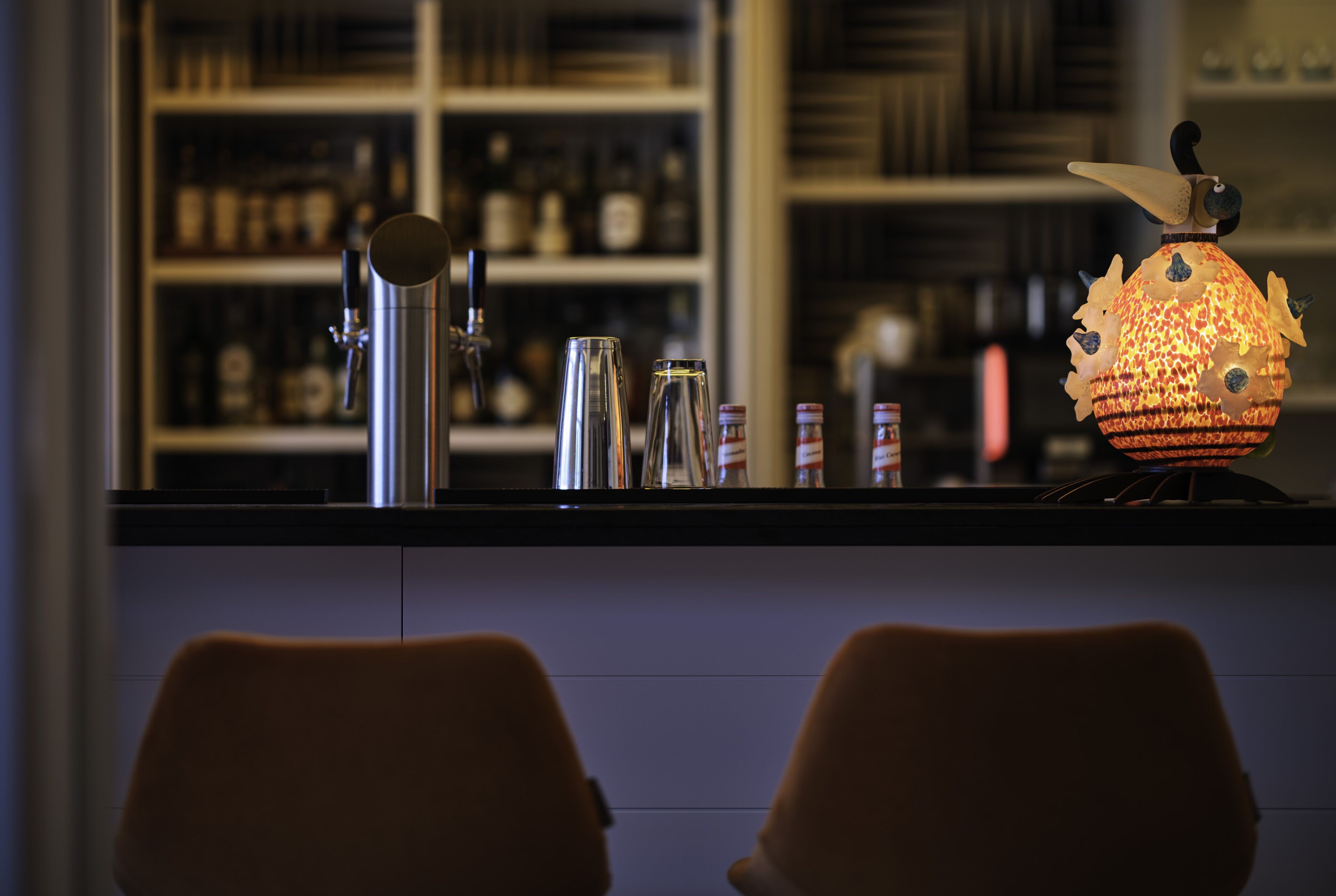 A bar area with a counter, shelves with bottles, and two chairs.