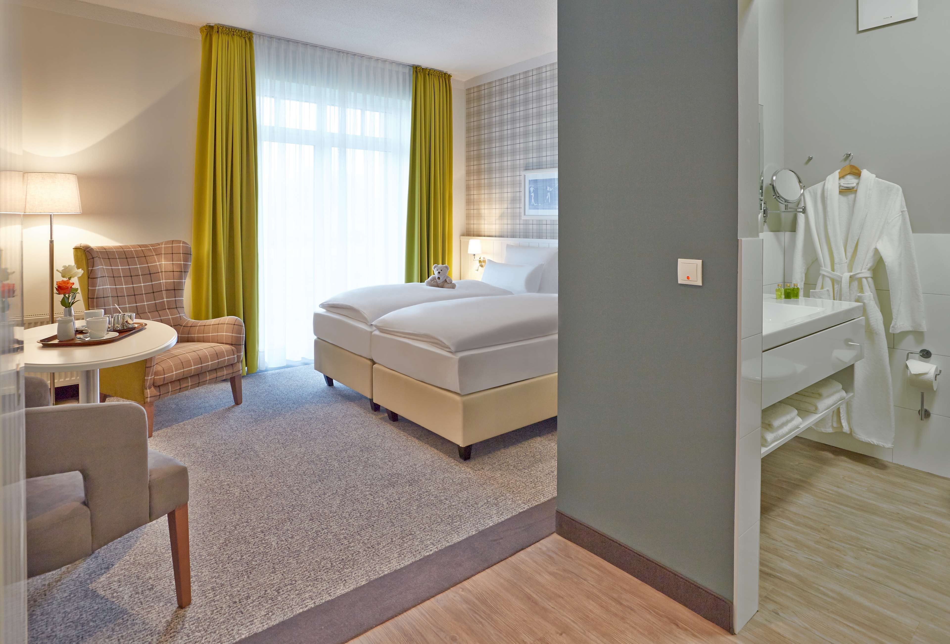 A hotel room with a bed, a desk with a chair, a small table with a lamp, a window with curtains, and a bathroom with a mirror and towel rack.