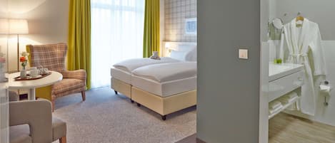 Ein Hotelzimmer mit Bett, Schreibtisch mit Stuhl, kleinem Tisch mit Lampe, Fenster mit Vorhängen sowie Bad mit Spiegel und Handtuchhalter.