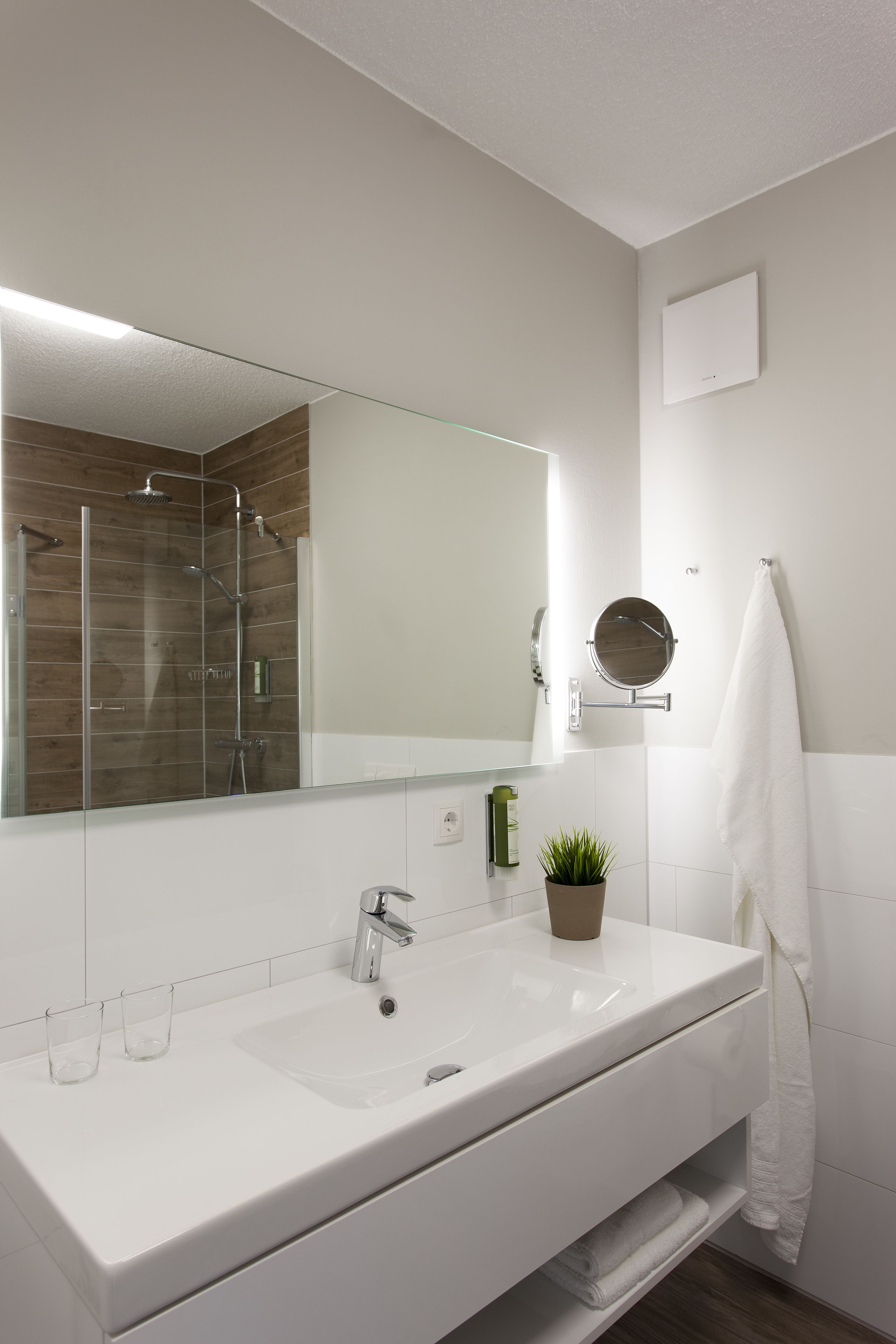 A modern bathroom with a large mirror, a white sink, a shower, and a towel hanging on the wall.