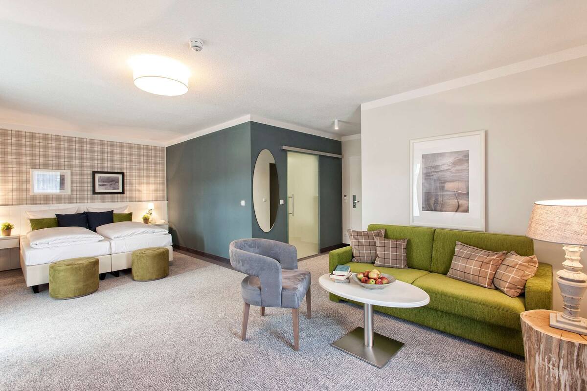 Una sala de estar moderna con un sofá verde, un sillón gris y una mesa de centro blanca. Hay un sofá blanco con cojines verdes y negros, y un pequeño banquillo verde. Un cuadro enmarcado cuelga de la pared, y una lámpara está sobre una mesita de madera.