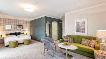 Un salon moderne avec un canapé vert, un fauteuil gris et une table basse blanche. On y trouve un canapé blanc avec des coussins verts et noirs, ainsi qu’un petit pouf vert. Un tableau encadré est accroché au mur, et une lampe est posée sur une table d’appoint en bois.