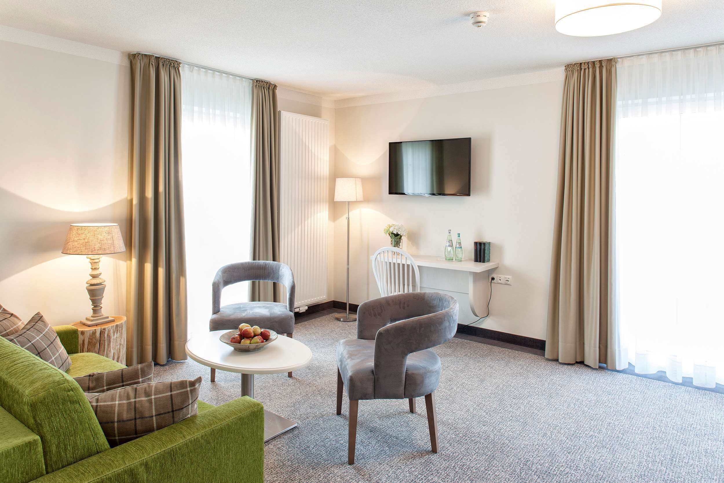 A modern hotel room with a green sofa, a grey armchair, a small round table with fruit, a lamp, a flat-screen TV, and a radiator.