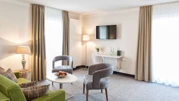 A modern hotel room with a green sofa, a grey armchair, a small round table with fruit, a lamp, a flat-screen TV, and a radiator.