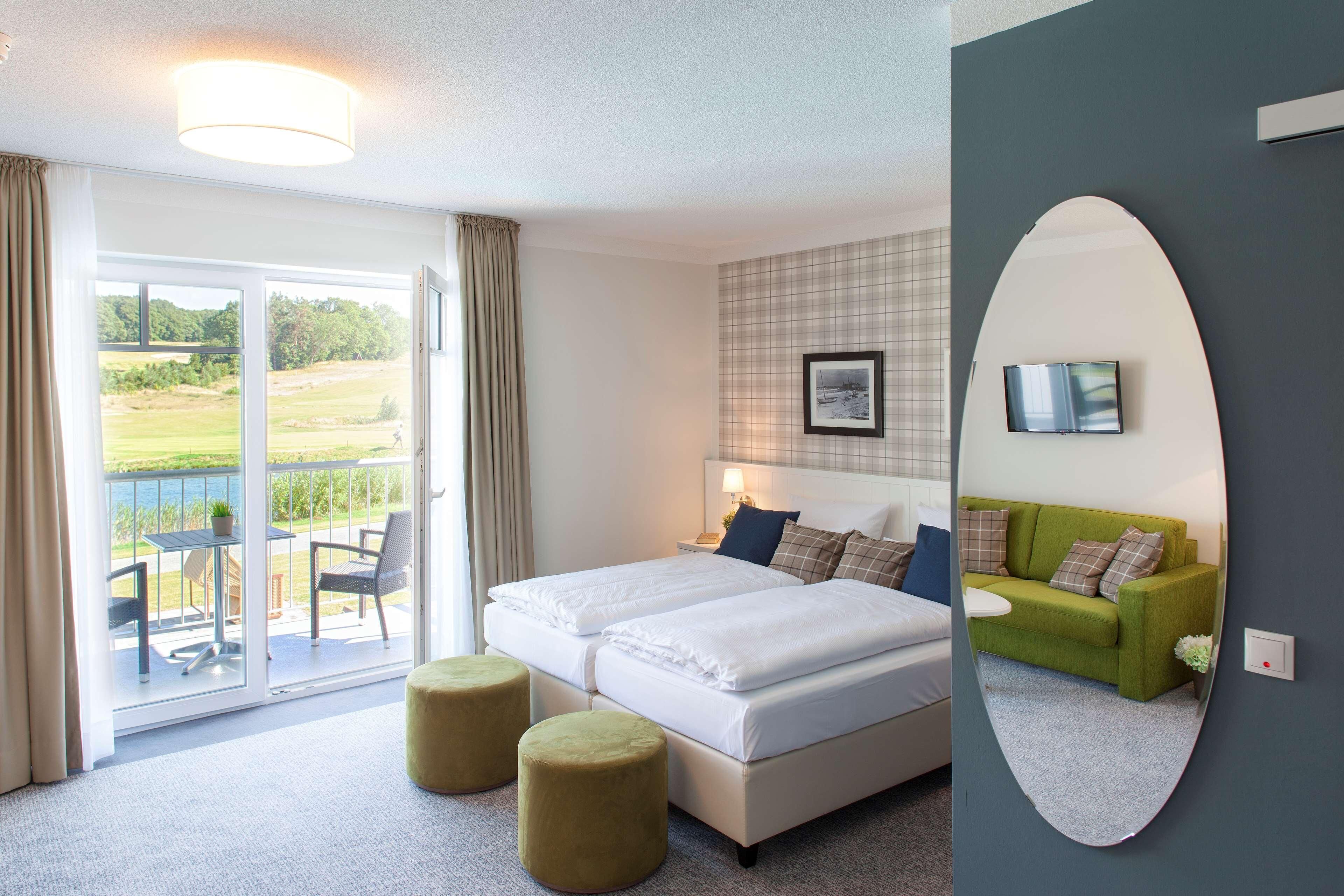 Habitación de hotel con cama, sofá verde, zona de comedor con mesa y sillas, y balcón con vistas.
