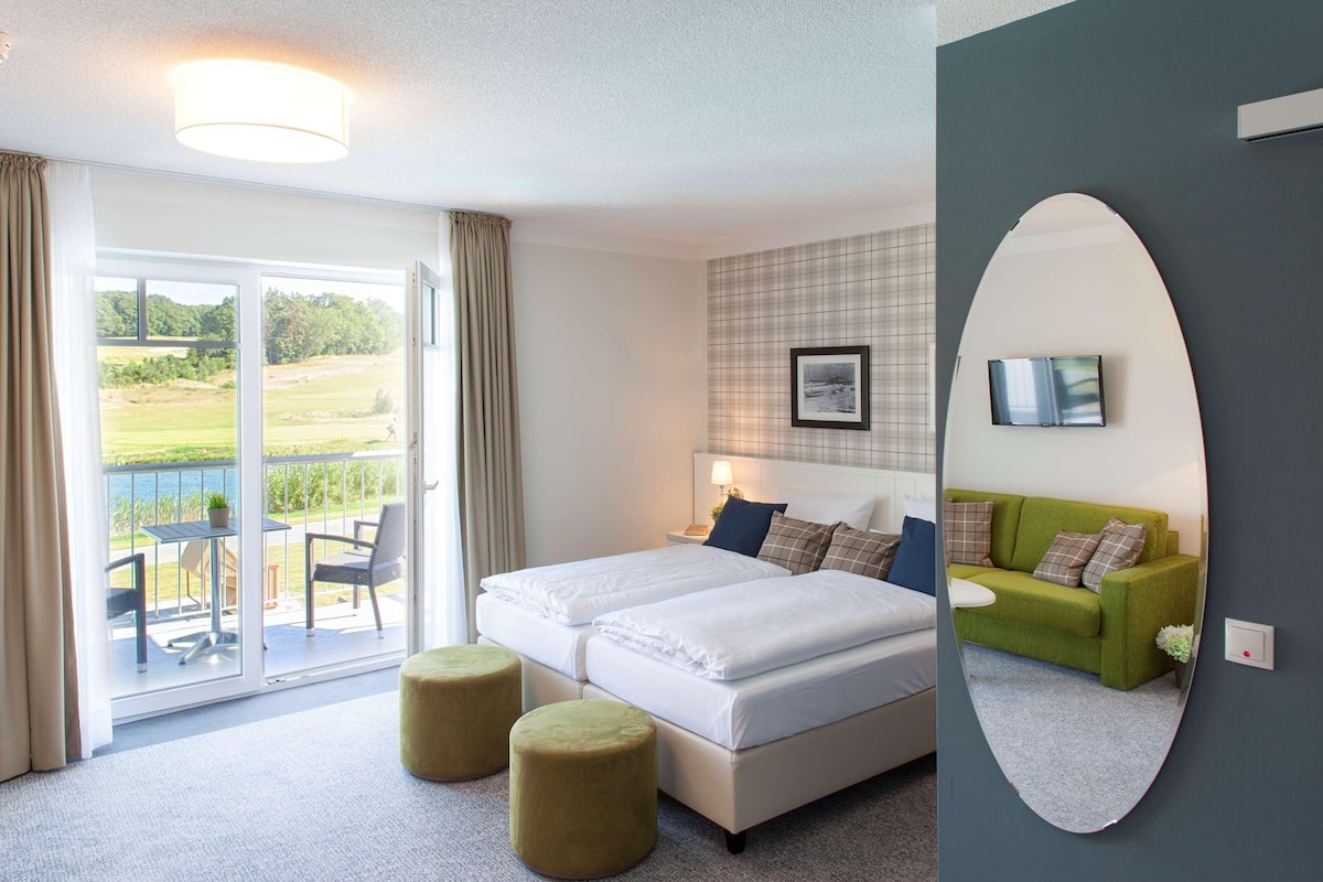Habitación de hotel con cama, sofá verde, zona de comedor con mesa y sillas, y balcón con vistas.