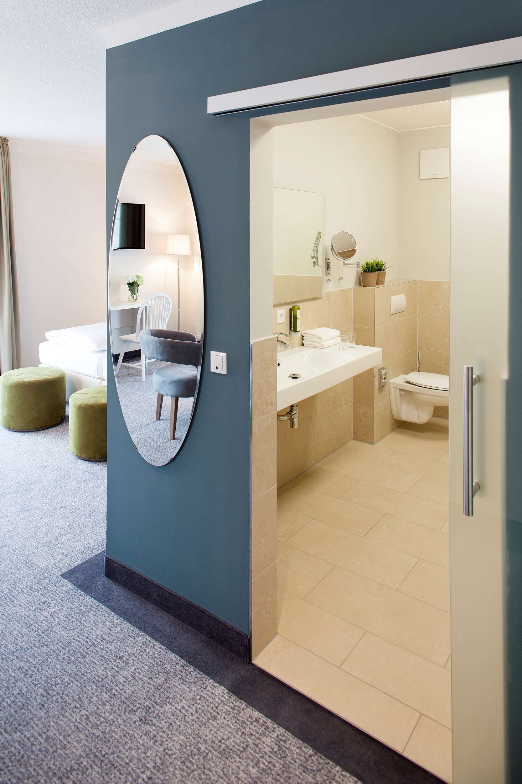 A modern hotel room with a large mirror, a bed, a desk, and a bathroom visible through an open door.