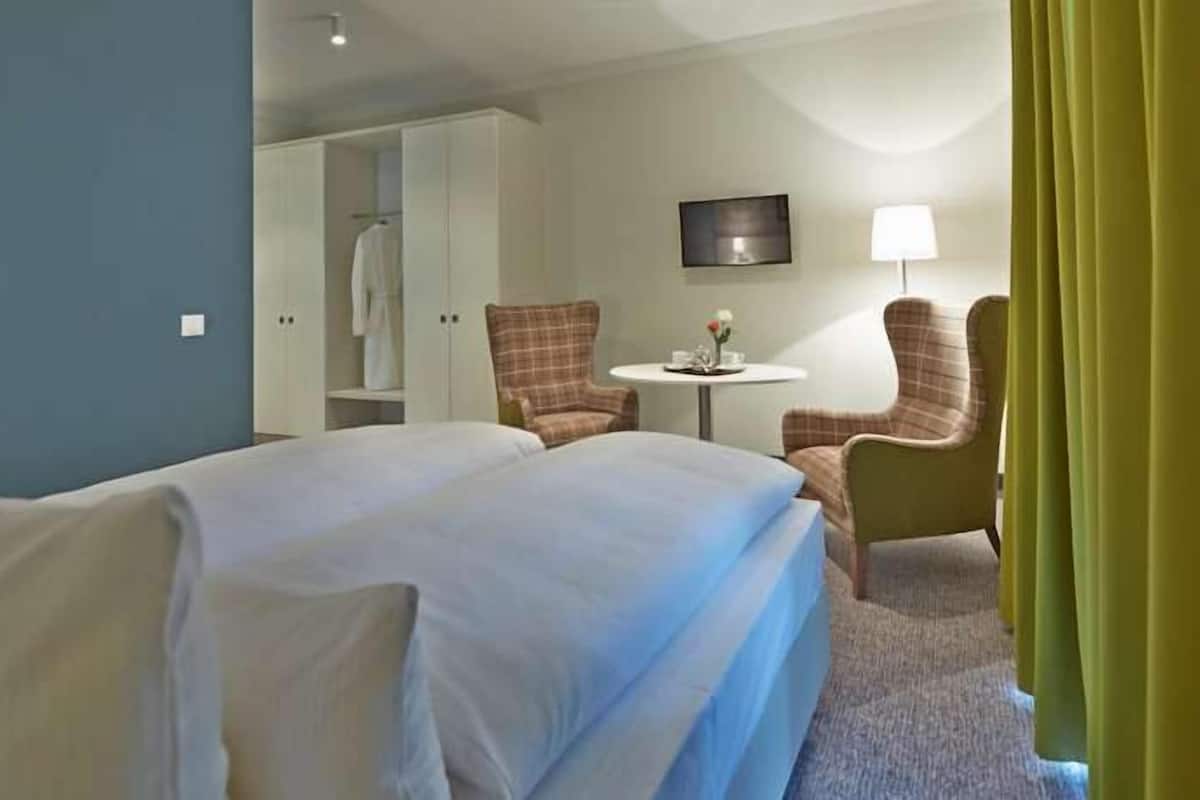 Una habitación de hotel con una cama, dos sillones, una mesita con un florero, un televisor en la pared y un armario.