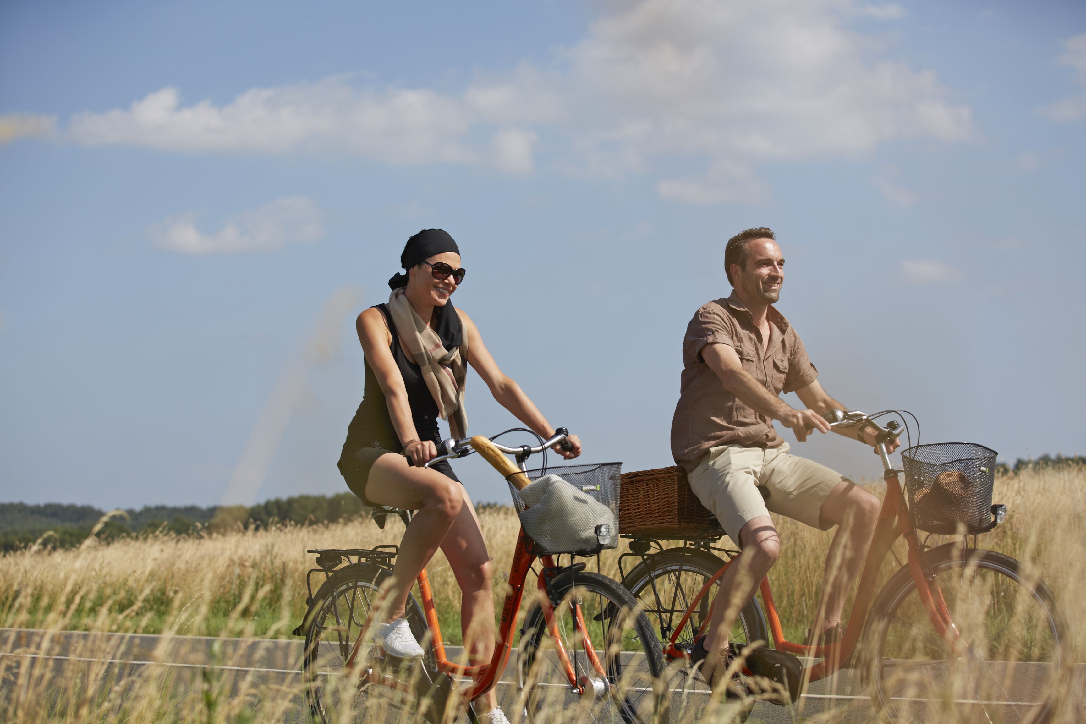 Two people riding bicycles with baskets, in a field with tall grass and a clear sky.