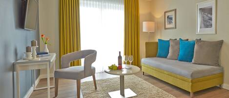A modern hotel room with a sofa, a chair, a small table with a wine bottle and glasses, a TV, and a window with yellow curtains.