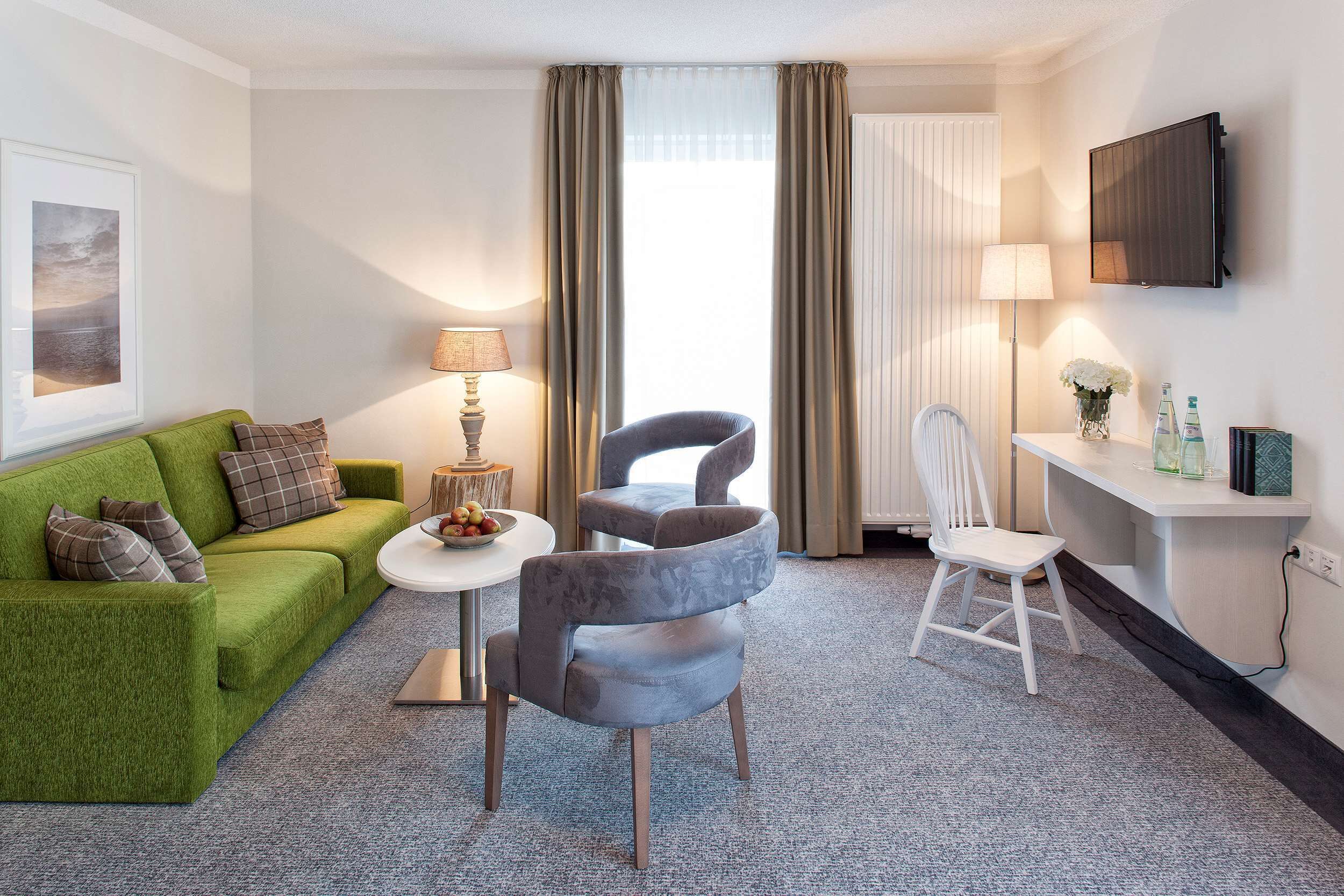 Hotellihuone, jossa on vihreä sohva, pyöreä pöytä lampun kanssa, moderni nojatuoli, valkoinen tuoli ja seinälle asennettu televisio.