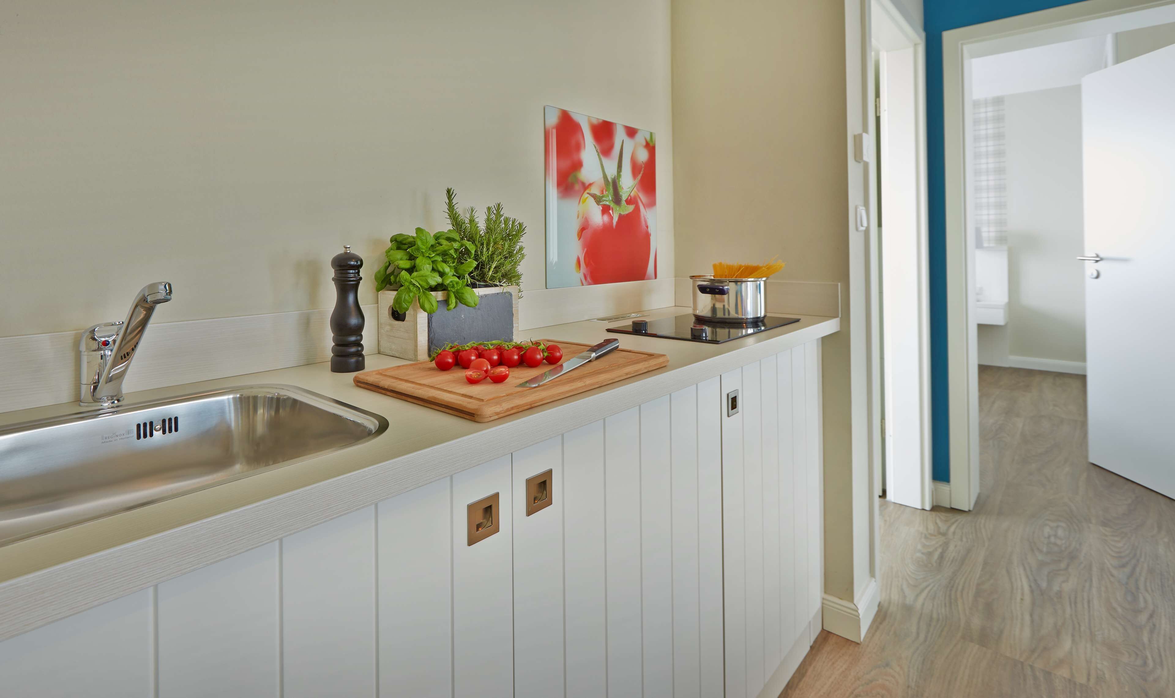 A kitchen with a stainless steel sink, a wooden cutting board with tomatoes, a knife, a pepper mill, a potted basil plant, and a painting of tomatoes on the wall.
