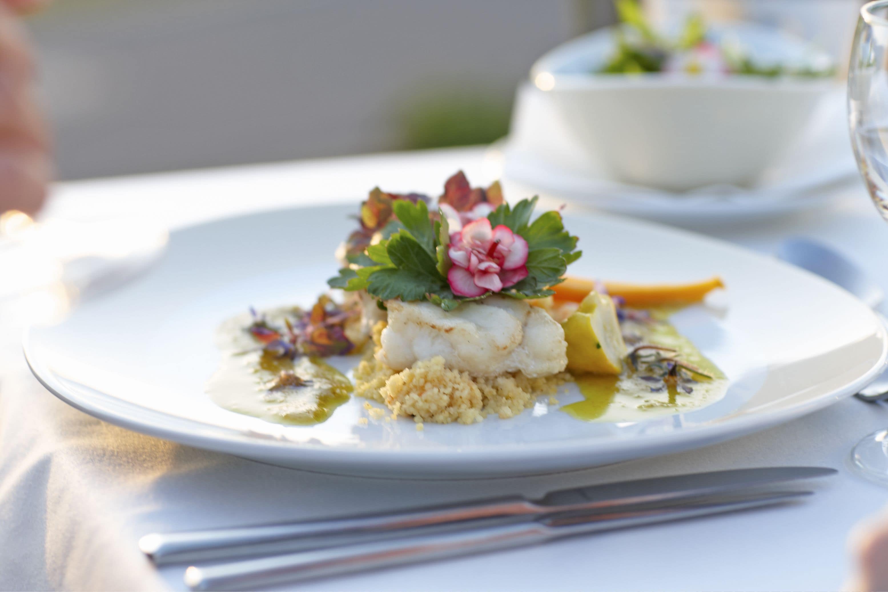 Un plato blanco con un plato de pescado, decorado con vegetales verdes y una flor rosa, sobre una mesa con mantel blanco.