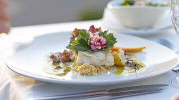 Un plato blanco con un plato de pescado, decorado con vegetales verdes y una flor rosa, sobre una mesa con mantel blanco.