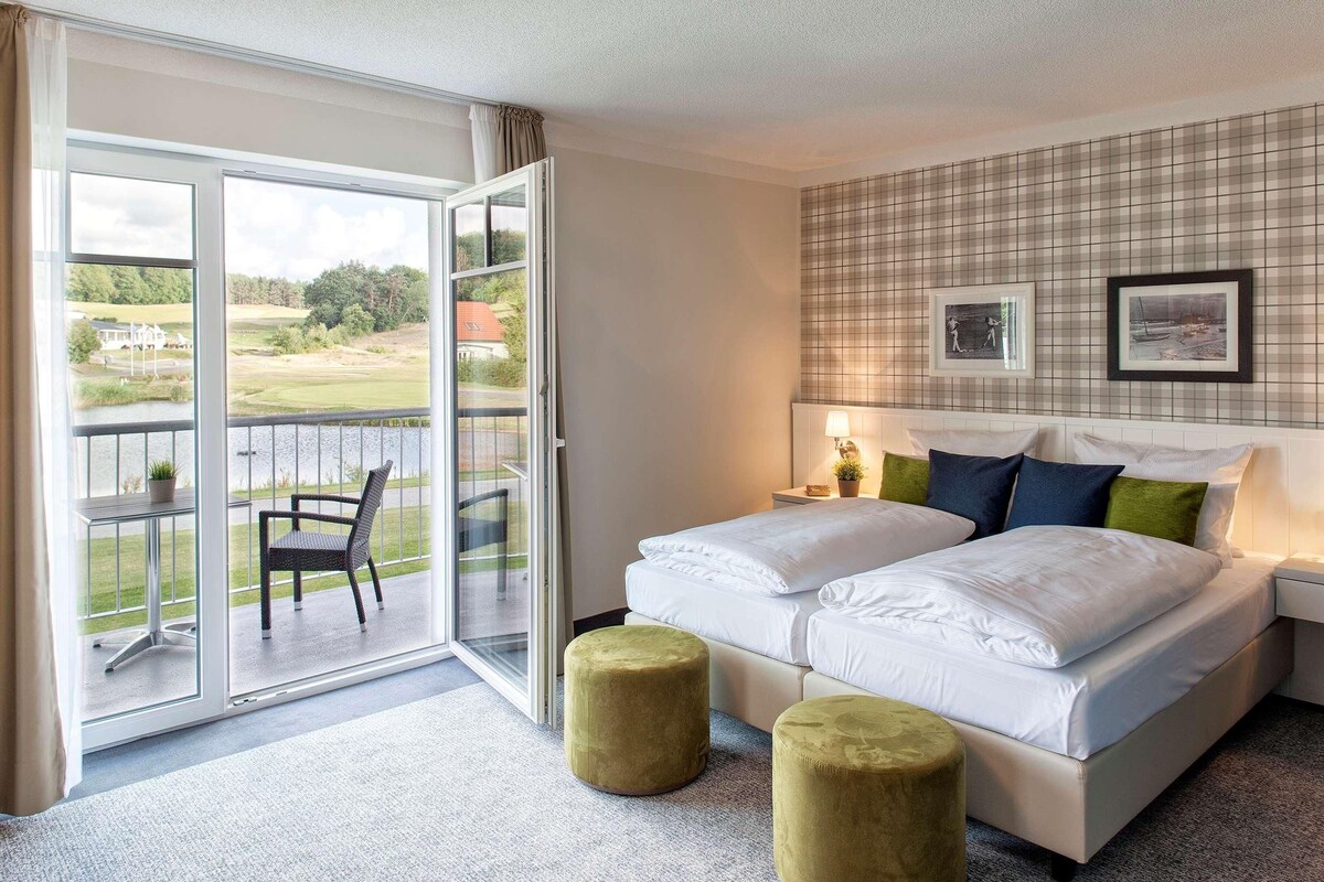 Habitación de hotel con cama, dos otomanas, una silla y vista a un campo de golf a través de una puerta corrediza de vidrio.