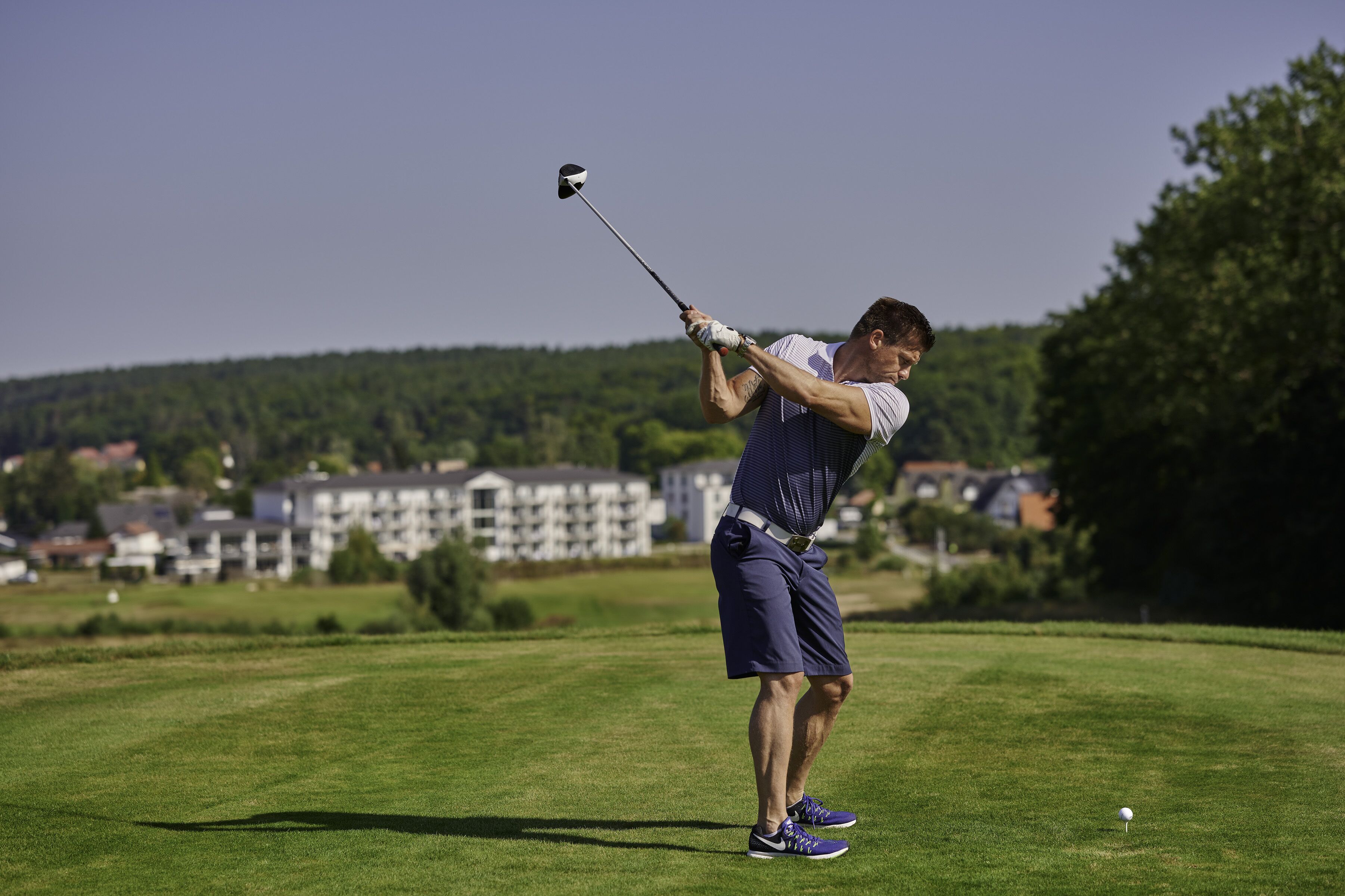 En person spiller golf på en velholdt bane med klar himmel og boliger i baggrunden.
