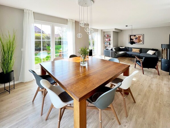A modern dining area with a wooden table and chairs, a potted plant, and a view of a garden through large windows.