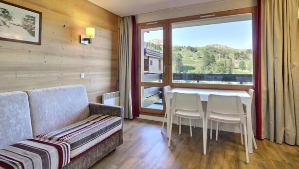 A room with a wooden wall, a sofa, a dining table with chairs, and a large window with a view of hills.