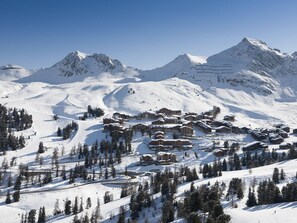 Estância de esqui nas montanhas coberta de neve, com vários edifícios em estilo chalé, pistas de esqui e céu claro.