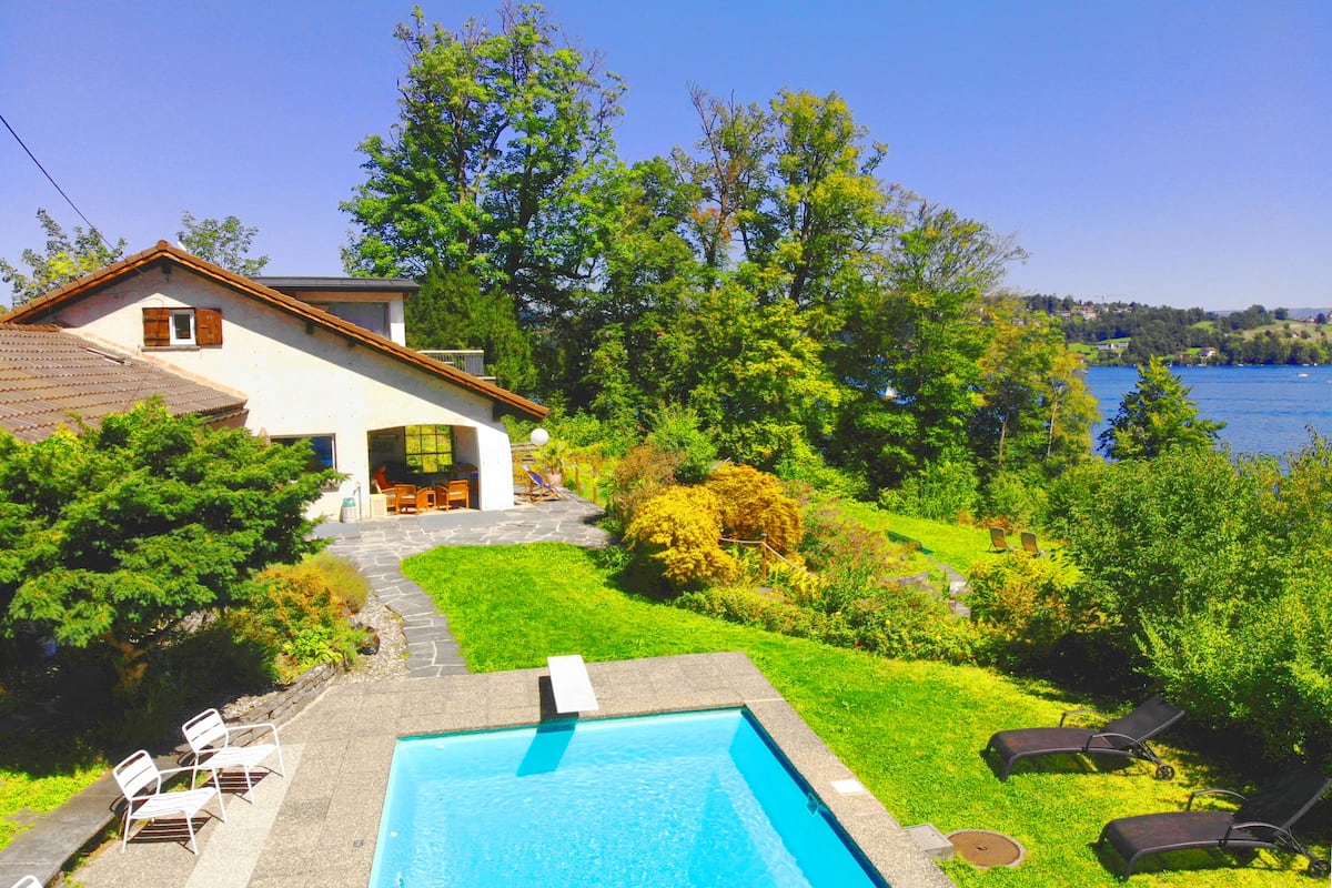 A swimming pool with lounge chairs, a house with a garden, and a view of a lake.