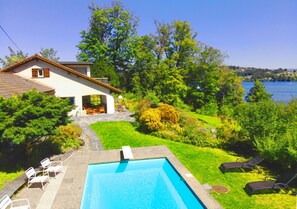 A swimming pool with lounge chairs, a house with a garden, and a view of a lake. - Lake Villa Lotus (Horw)