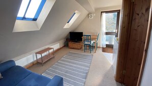 Ein gemütliches Schlafzimmer im Dachgeschoss mit Dachfenstern, einem Fernseher, einem Holztisch und einem blauen Sofa.