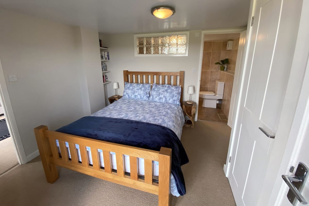 A bedroom with a wooden bed, a nightstand, a bedside table, a window with a decorative frame, and a bathroom with a toilet and sink.