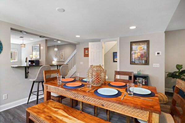 Un comedor con una mesa de madera dispuesta para seis personas, una escalera y un cuadro en la pared.