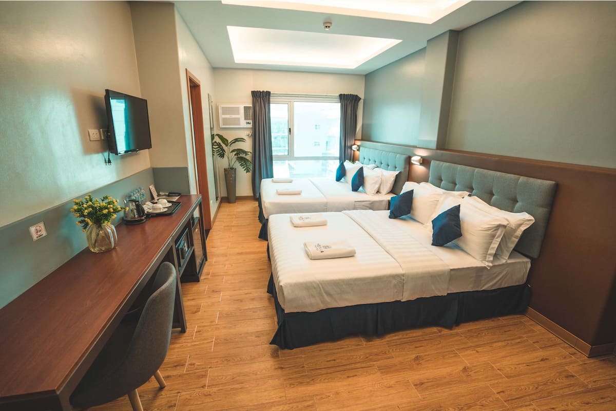 Quarto de hotel com duas camas, uma escrivaninha, televisão e janela com cortinas.