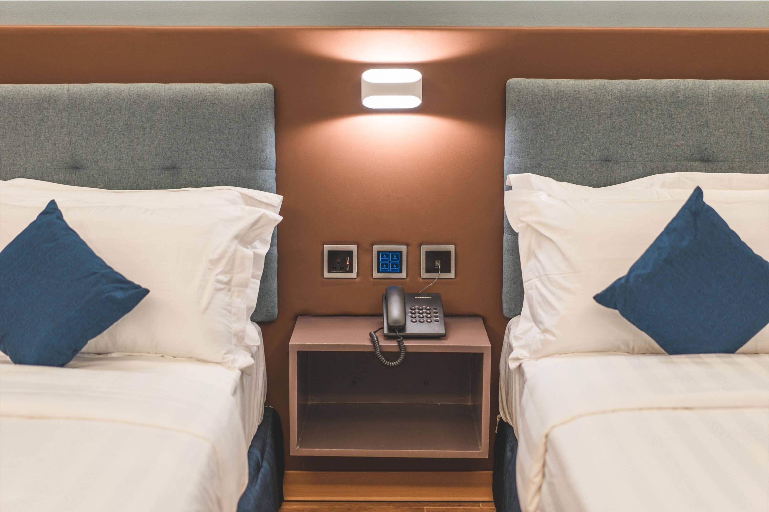 A hotel room with two beds, a nightstand with a phone, and a wall-mounted light.