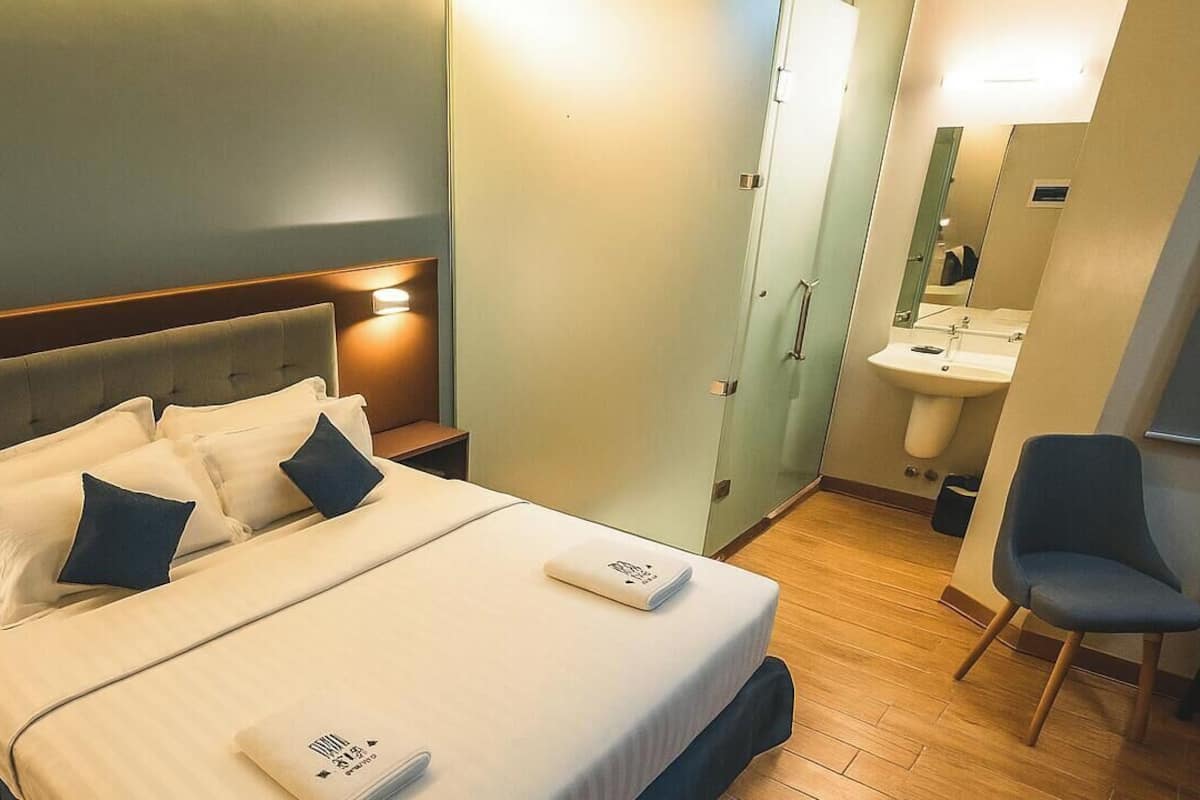 Quarto de hotel com cama, mesas de cabeceira, uma cadeira, banheiro com pia e espelho.